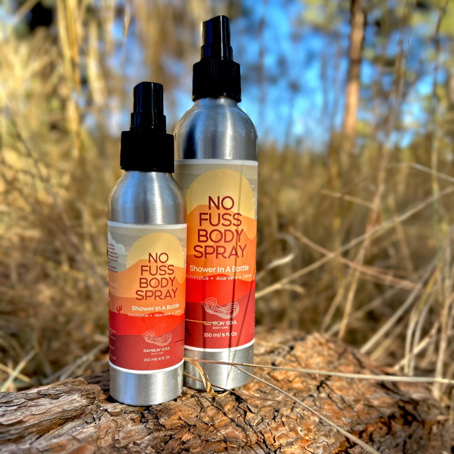 two silver aluminum spray bottles labeled 'NO FUSS BODY SPRAY' placed on a natural rock surface with green foliage in the background.